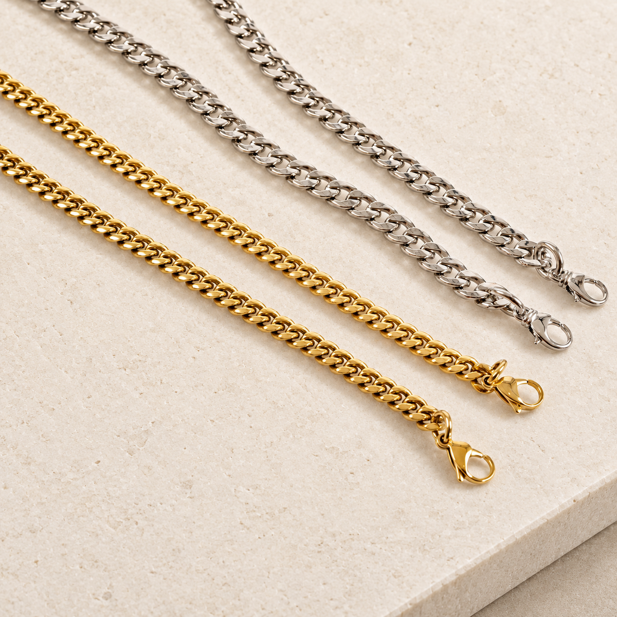 JaneFinds Cuban crossbody chains in gold and palladium with rounded swivel clasps on pale limestone.