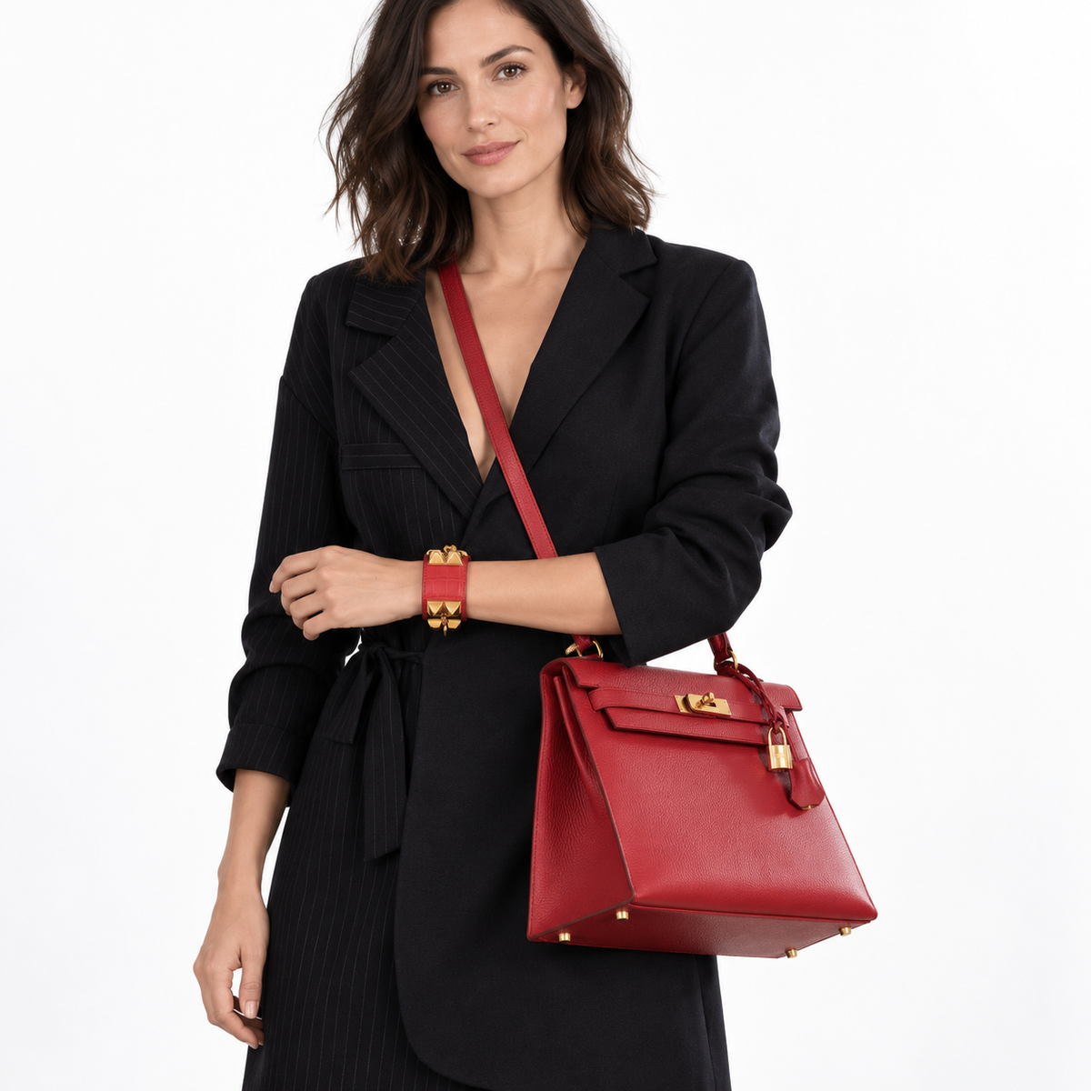 Hermès Kelly 32 Sellier Rouge Vif Ardennes with gold hardware worn crossbody by a model in a black tailored coat against a white studio background