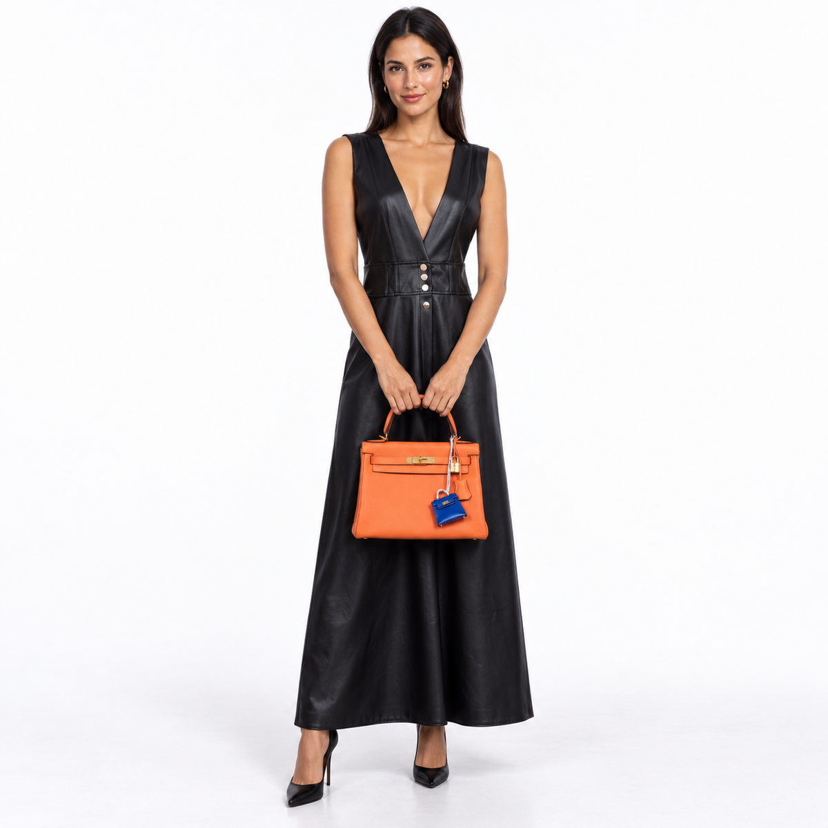 Hermès Kelly 28 Feu Orange Togo with gold hardware and blue miniature Kelly charm carried by a woman in a black dress in a white studio
