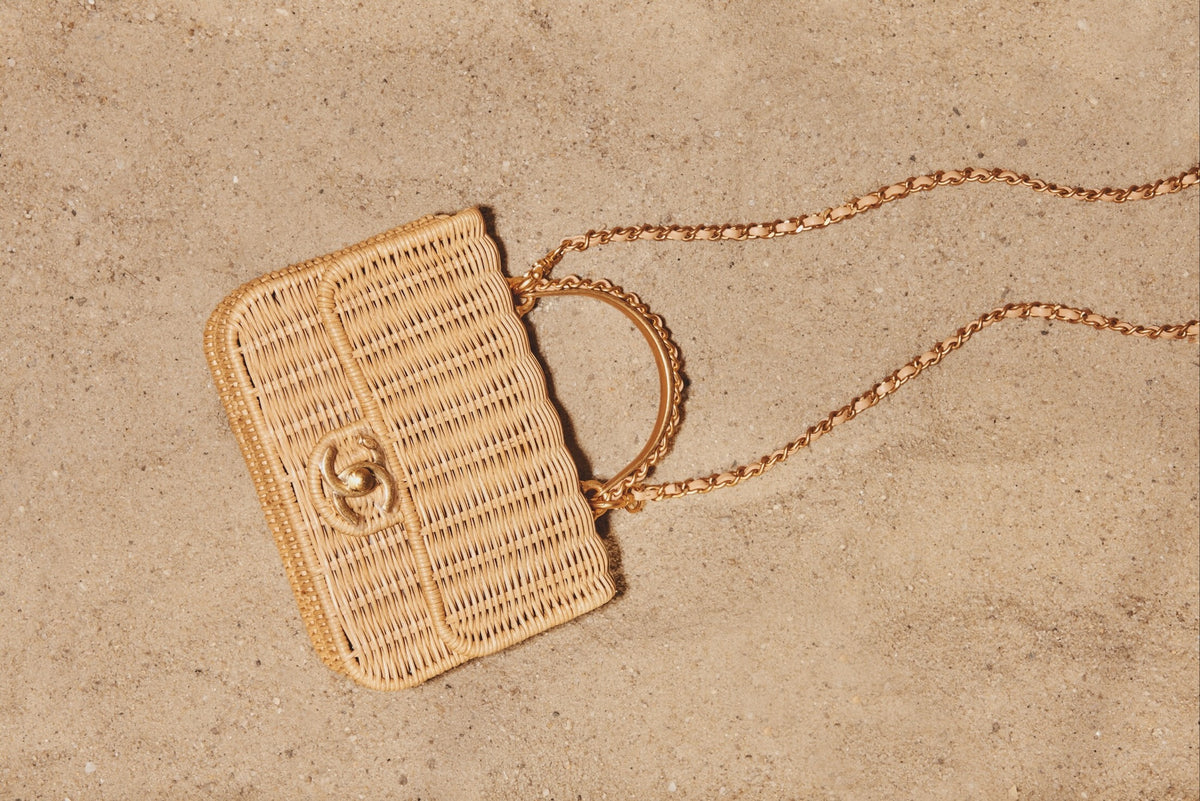 Chanel Mini Flap Bag with Top Handle Beige Braided Rattan Gold-Tone Hardware, lying on sand with its chain strap