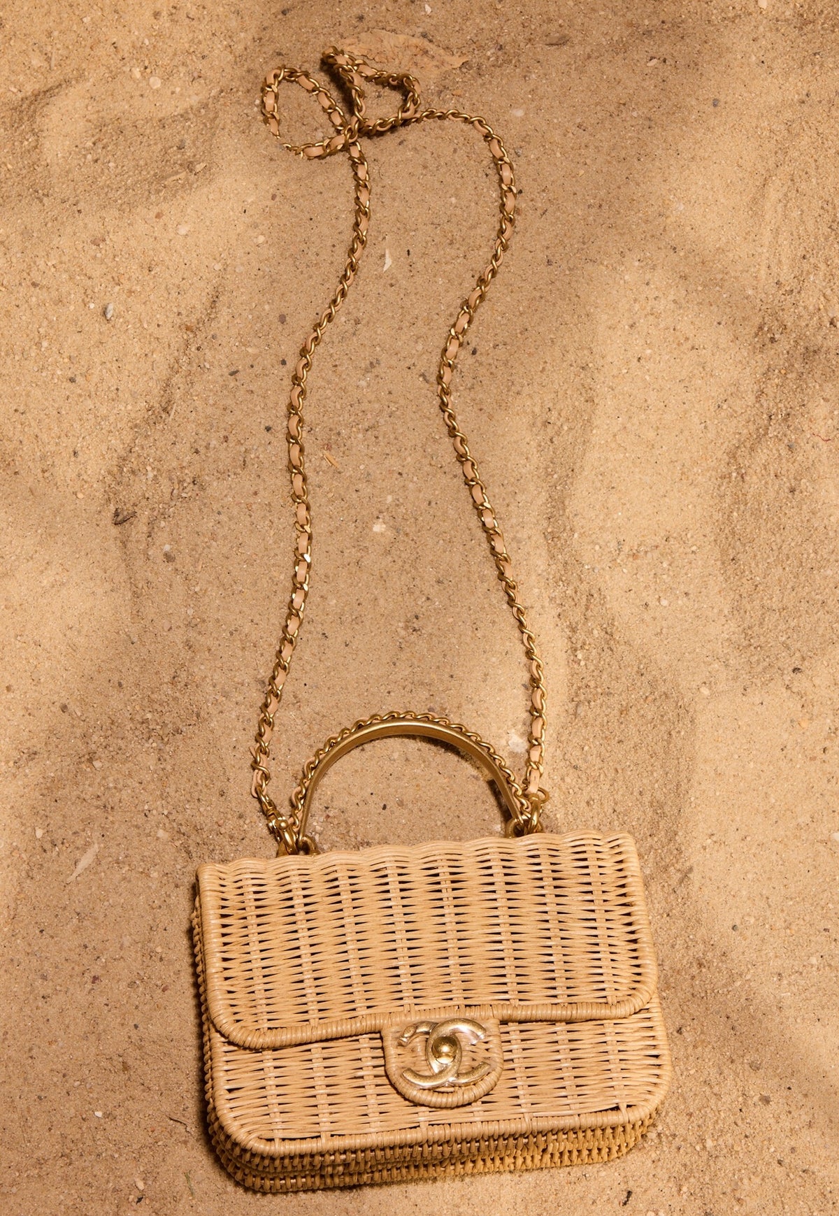 Chanel Mini Flap Bag with Top Handle Beige Braided Rattan Gold-Tone Hardware, lying on sand with its chain strap, top view