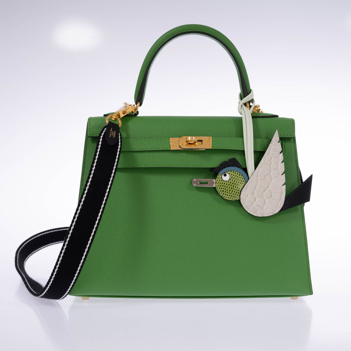 Hermès Birdy bag charm with a pale alligator wing and green lizard head, attached to a green Kelly beside a black shoulder strap, both sold separately.