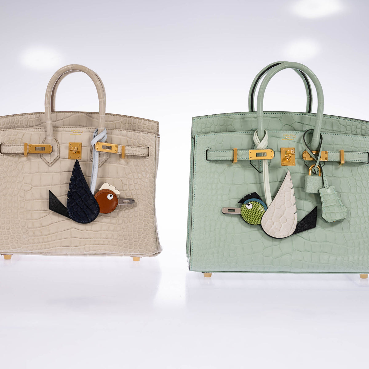 Hermès Birdy bag charm in Béton, Noir and Pelouse on a pale green exotic-leather Birkin at right, beside a cream Birkin with a different Birdy charm, all sold separately.