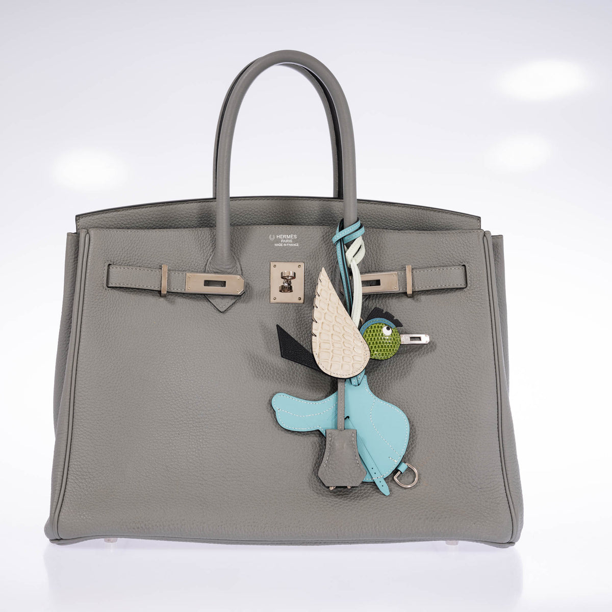 Hermès Birdy bag charm in Béton, Noir and Pelouse on a gray Birkin, beside a pale blue saddle-shaped accessory and the bag's clochette, all sold separately.