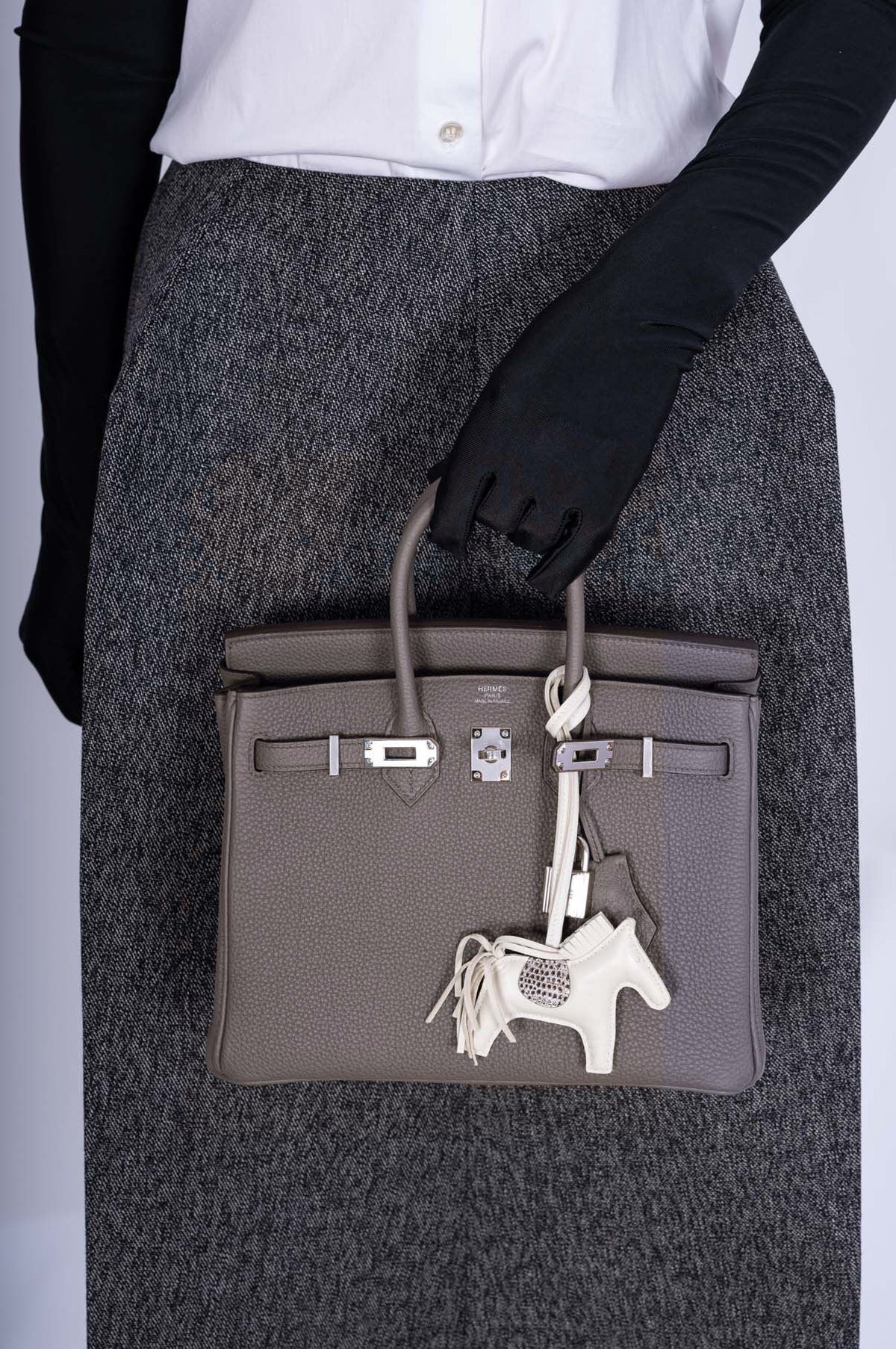 Hermès Birkin 25 Étain Togo Palladium Hardware, close-up carried by a model in black gloves and a gray skirt, with an ivory horse charm, sold separately.