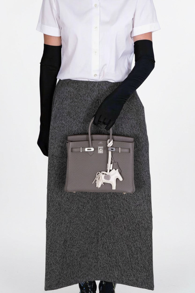 Hermès Birkin 25 Étain Togo Palladium Hardware, carried by a model in a white shirt, long gray skirt and black gloves, with an ivory horse charm, sold separately.