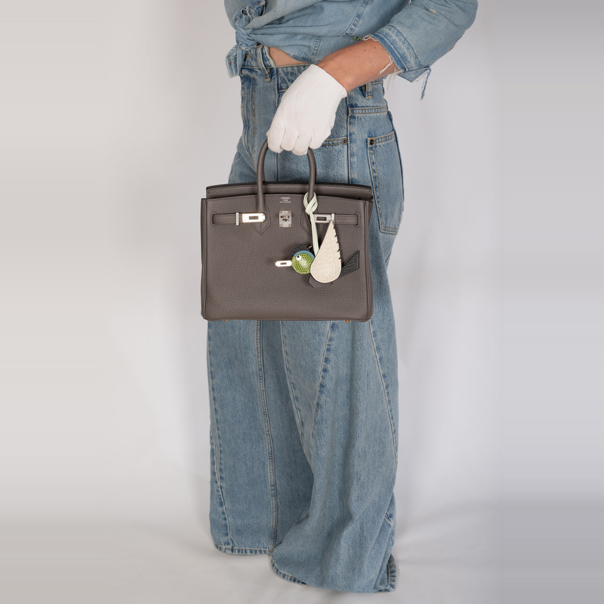 Hermès Birkin 25 Étain Togo Palladium Hardware, carried by a model in denim and white gloves with a Hermès Birdy charm, sold separately.