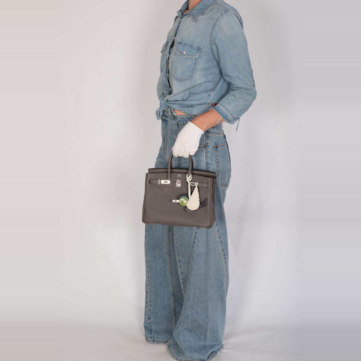 Hermès Birkin 25 Étain Togo Palladium Hardware, full-length view carried by a model in denim and white gloves with a Hermès Birdy charm, sold separately.