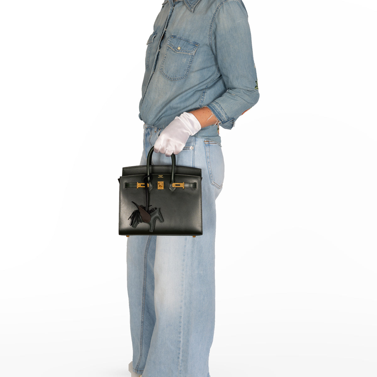 Hermès Birkin 25 Sellier Vert Foncé Box Calf Gold Hardware, carried at the hip by a model in denim and white gloves in a white studio, with a winged horse charm sold separately.