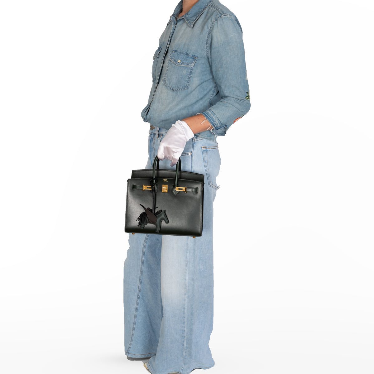 Hermès Birkin 25 Sellier Vert Foncé Box Calf Gold Hardware, hand-carried by a model in denim in a white studio, with a winged horse charm sold separately.