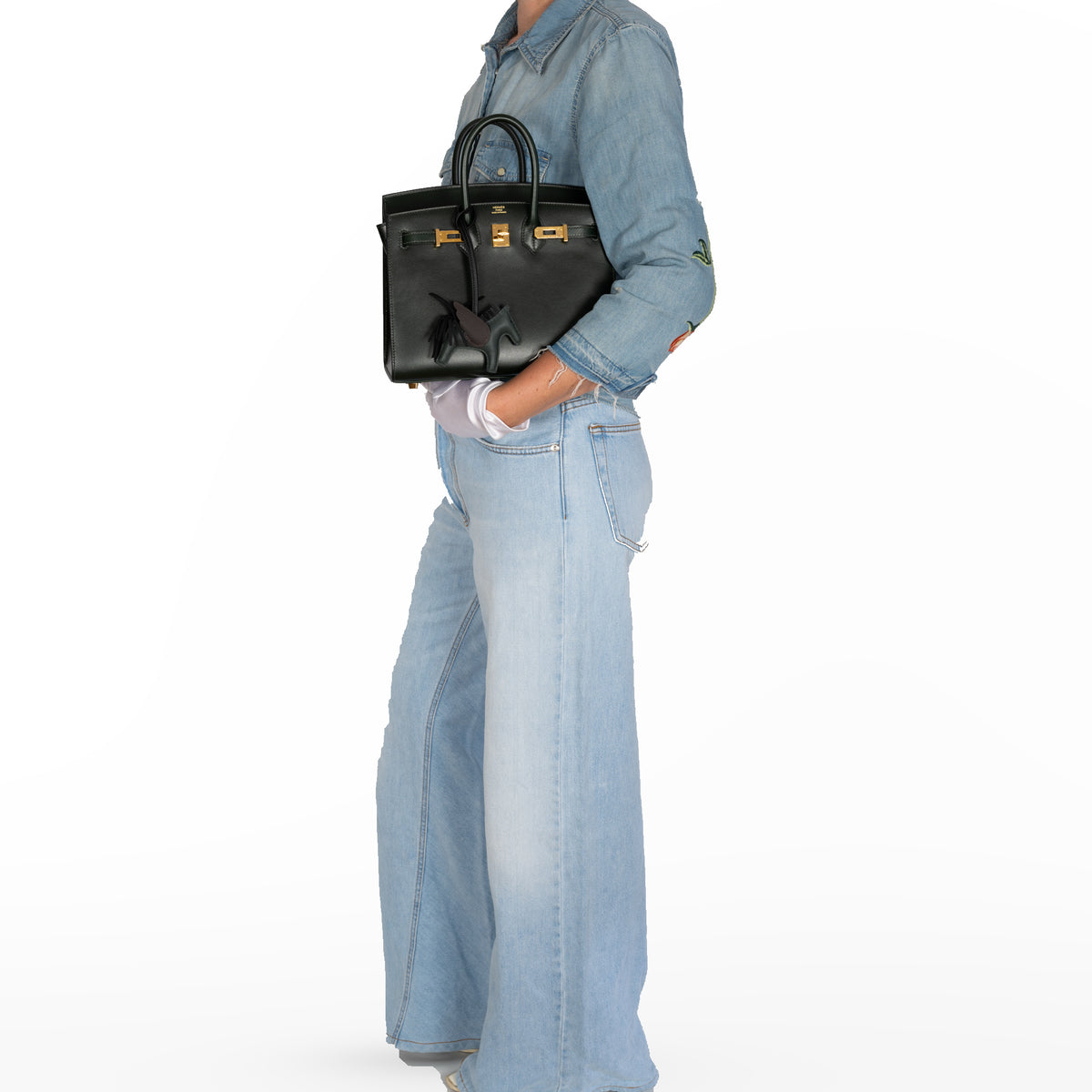 Hermès Birkin 25 Sellier Vert Foncé Box Calf Gold Hardware, held against a denim-clad model's body in a white studio, with a winged horse charm sold separately.