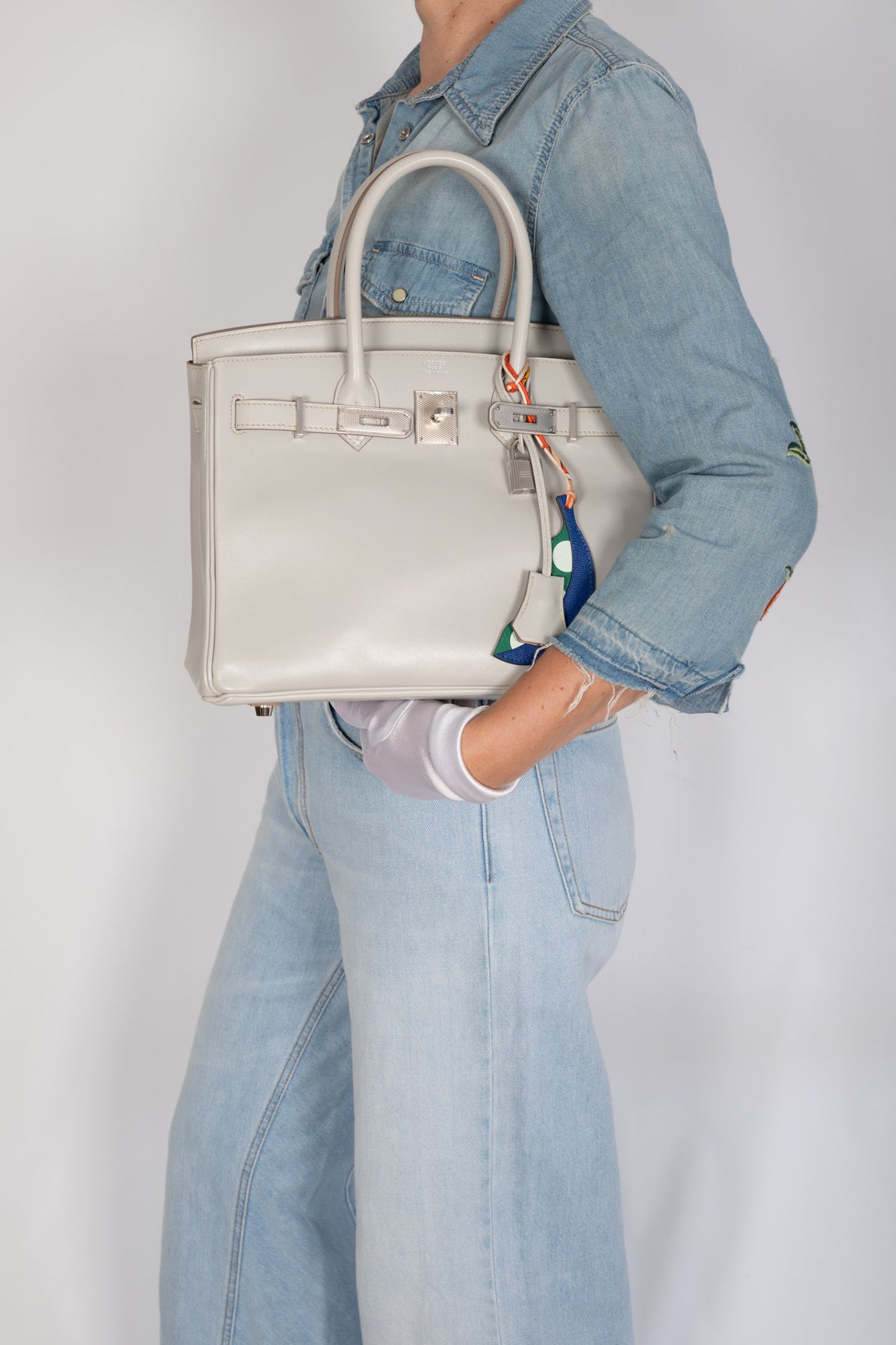 Hermès Birkin 30 Gris Perle Box Calf Guilloche Palladium Hardware, carried on the forearm by a model in denim, styled with a blue and green pea pod charm, sold separately