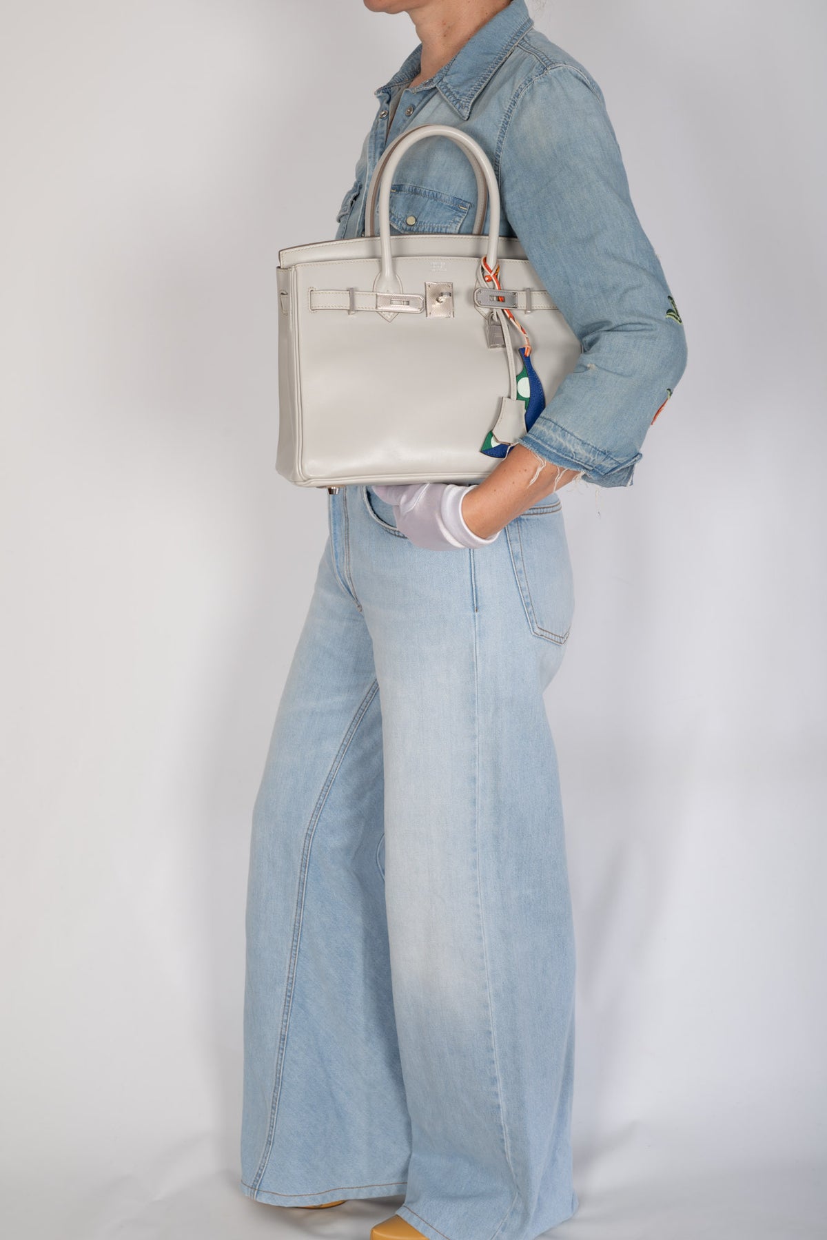 Hermès Birkin 30 Gris Perle Box Calf Guilloche Palladium Hardware, full-length view carried on the forearm by a model in denim, styled with a blue and green pea pod charm, sold separately