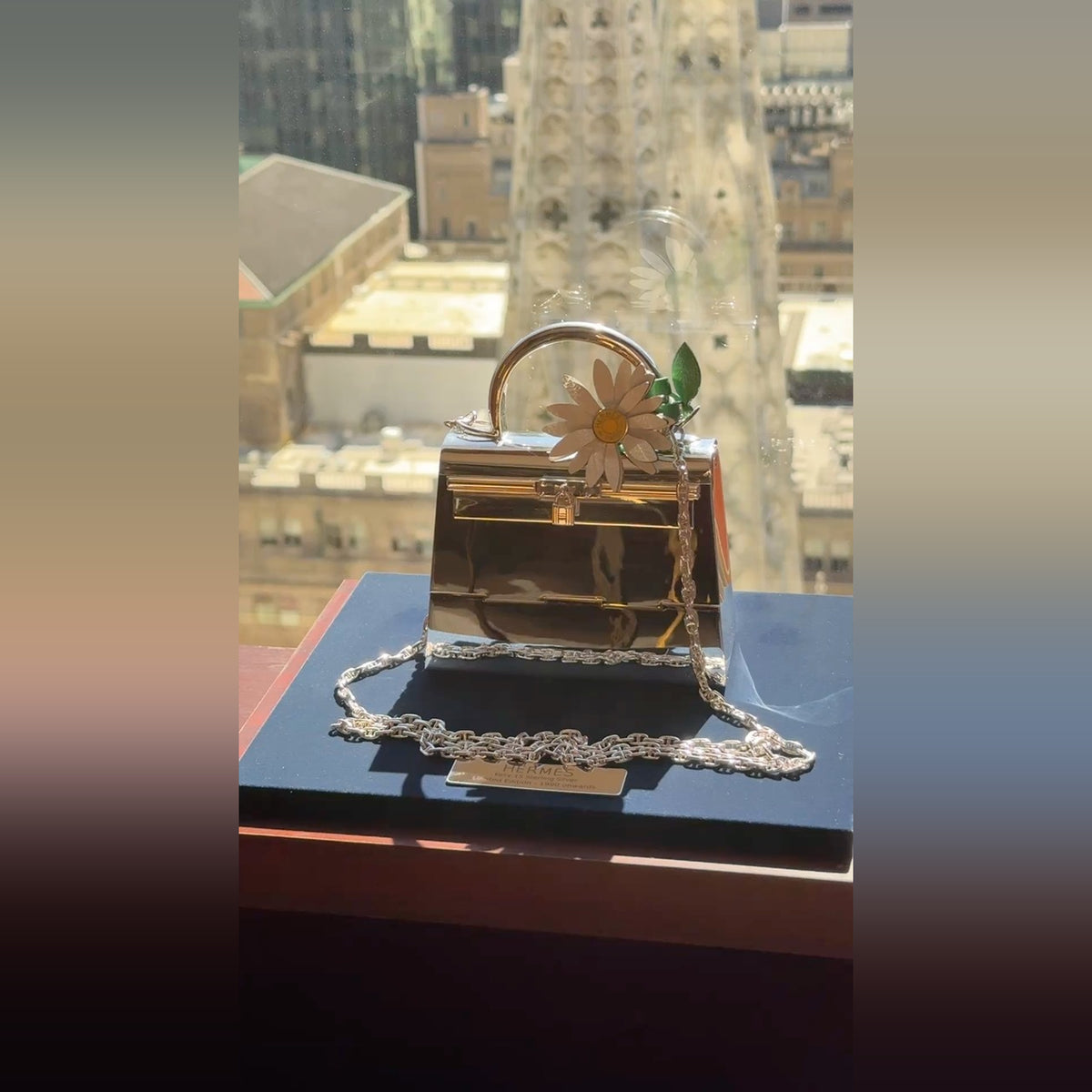 Hermès Kelly 15 Sterling Silver Chaîne d'Ancre Strap, on its stand by a window overlooking the city, shown with a marguerite charm, charm sold separately