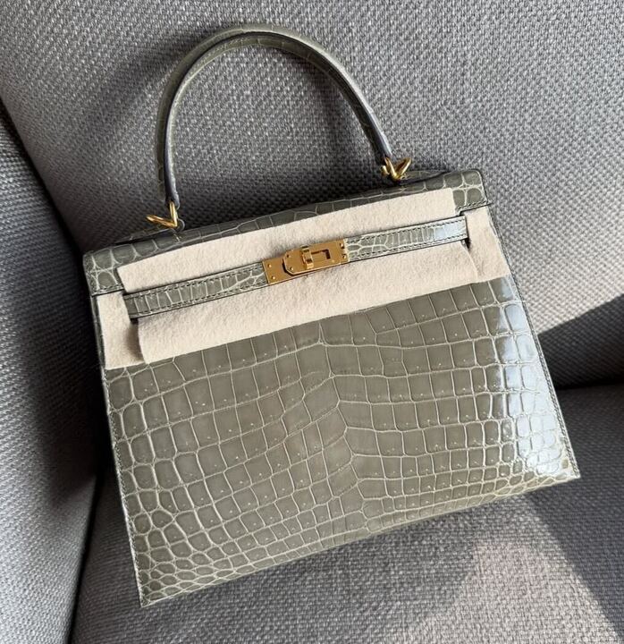 Hermès Kelly 25 Sellier Gris Tourterelle Shiny Niloticus Gold Hardware, front view on a sofa with protective felt