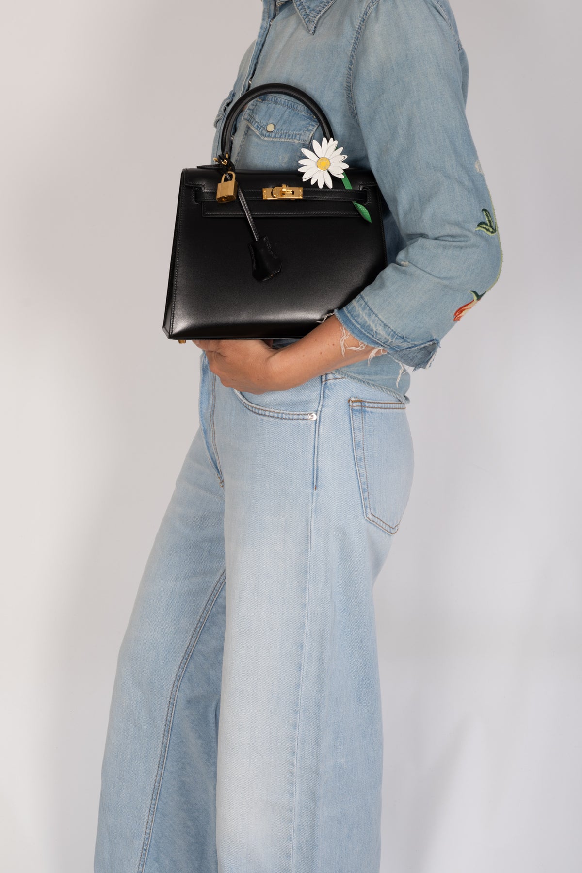Hermès Kelly 25 Sellier in Noir Box calf with gold hardware, carried by a model with the Hermès Marguerite Daisy Charm, sold separately.