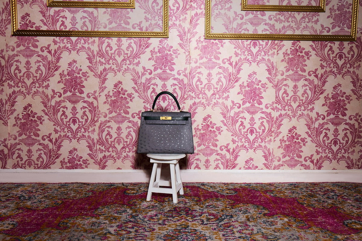 Hermès Kelly 28 Sellier HSS Gris Agate/Vert Titien Ostrich Brushed Gold Hardware, on a white stool against damask wallpaper