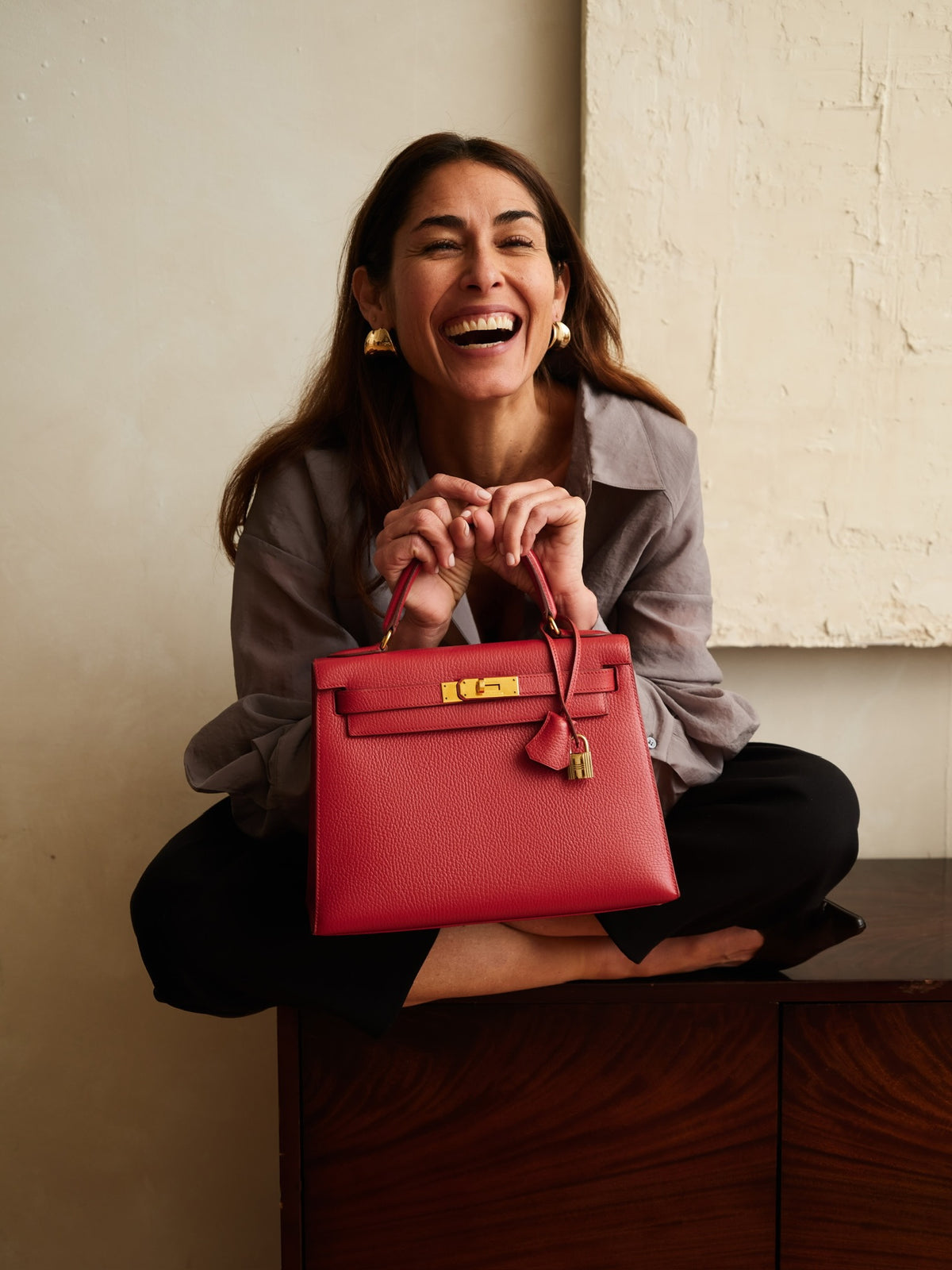 Hermès Kelly 28 Sellier Rouge Vif Ardennes Gold Hardware, held by a laughing model seated on a sideboard