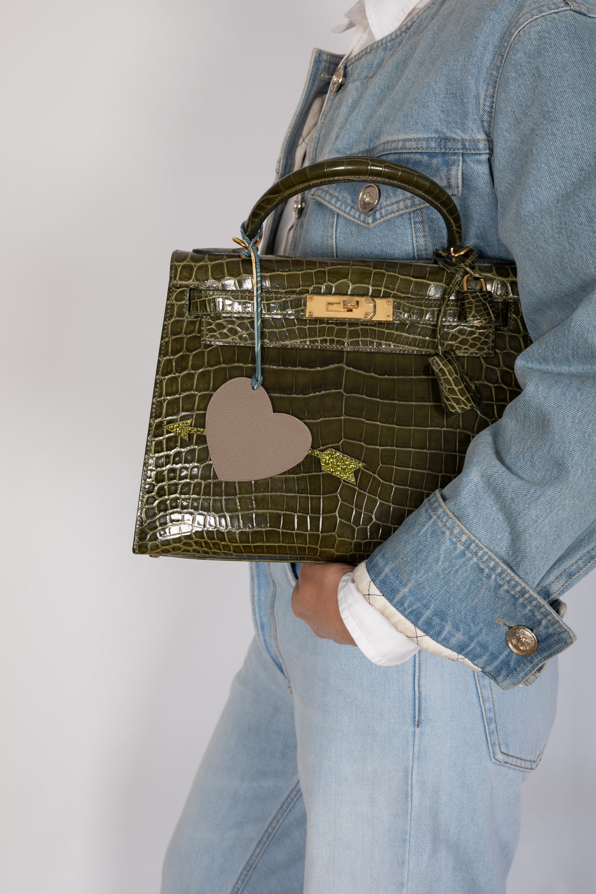 Hermès Kelly 28 Sellier Vert Véronèse shiny Niloticus crocodile, carried under the arm by a model in a denim jacket, with a heart charm, sold separately.