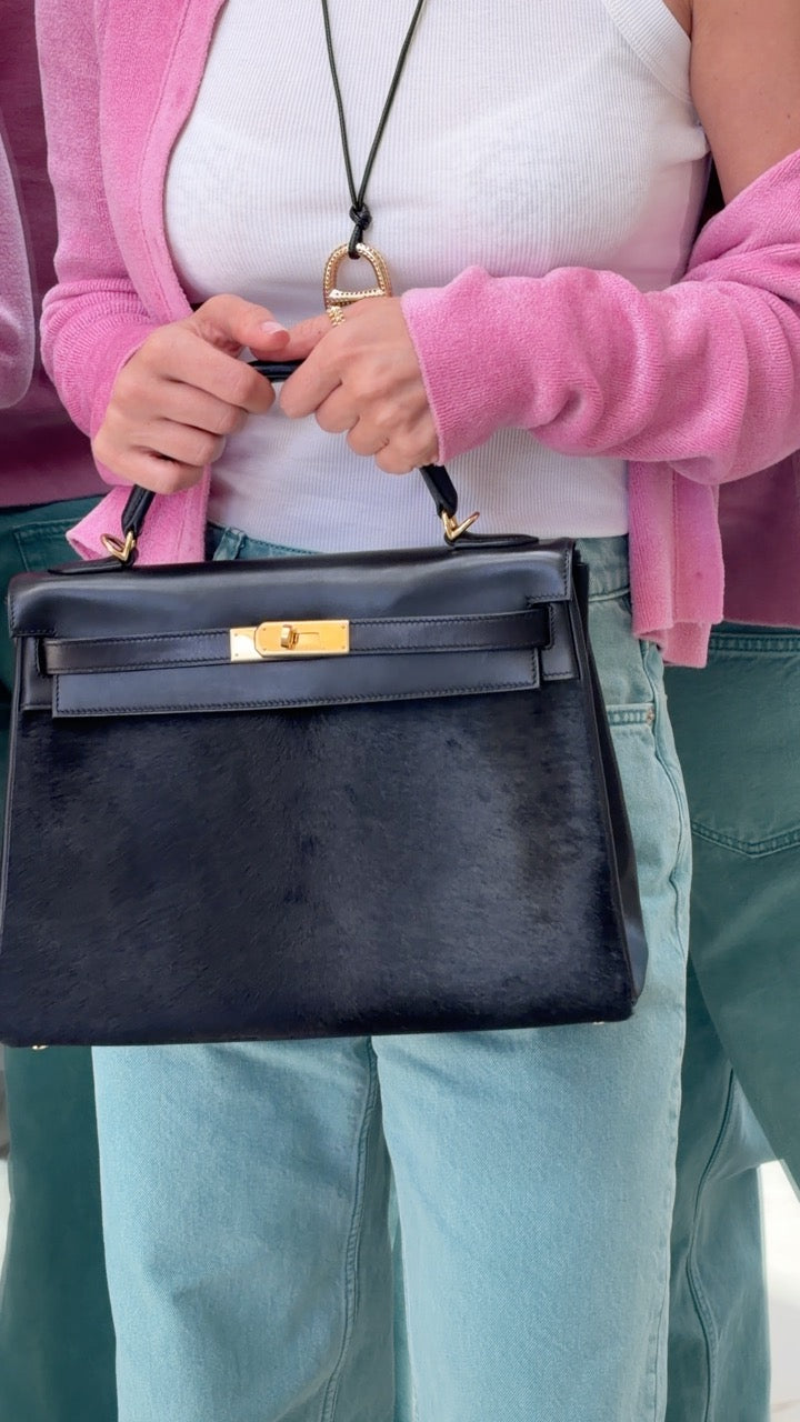 Hermès Kelly 32 Noir Box leather and pony skin with Gold hardware, held by its top handle with a pink cardigan and jeans.