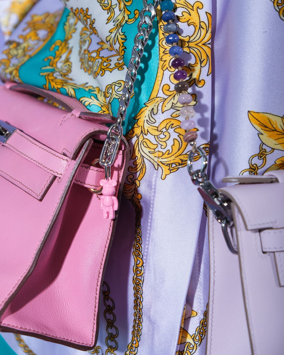 Hermès Kelly Pochette 5P Swift with Palladium Hardware, worn by a model beside a lilac Kelly Pochette, close view with a bear charm and a beaded strap