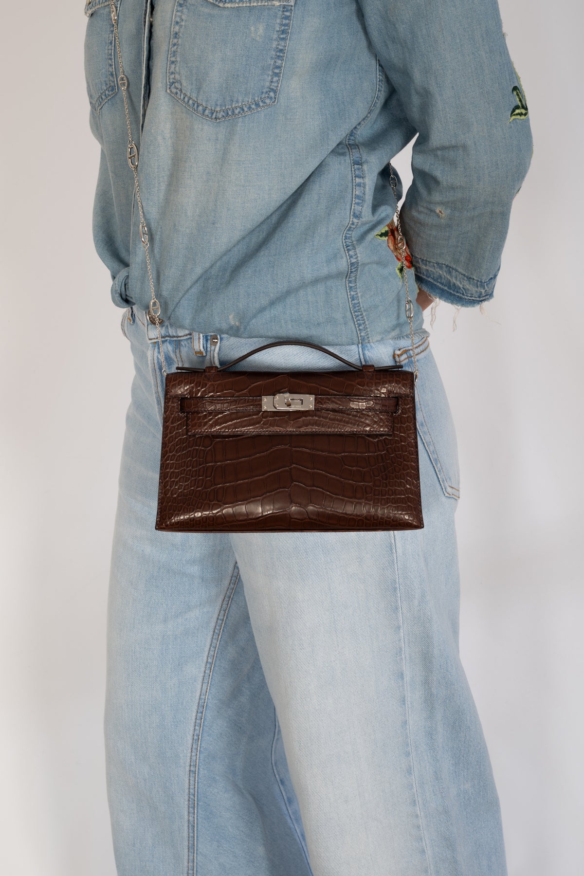 Hermès Kelly Pochette in Havane matte alligator, worn crossbody on a silver chain with denim.