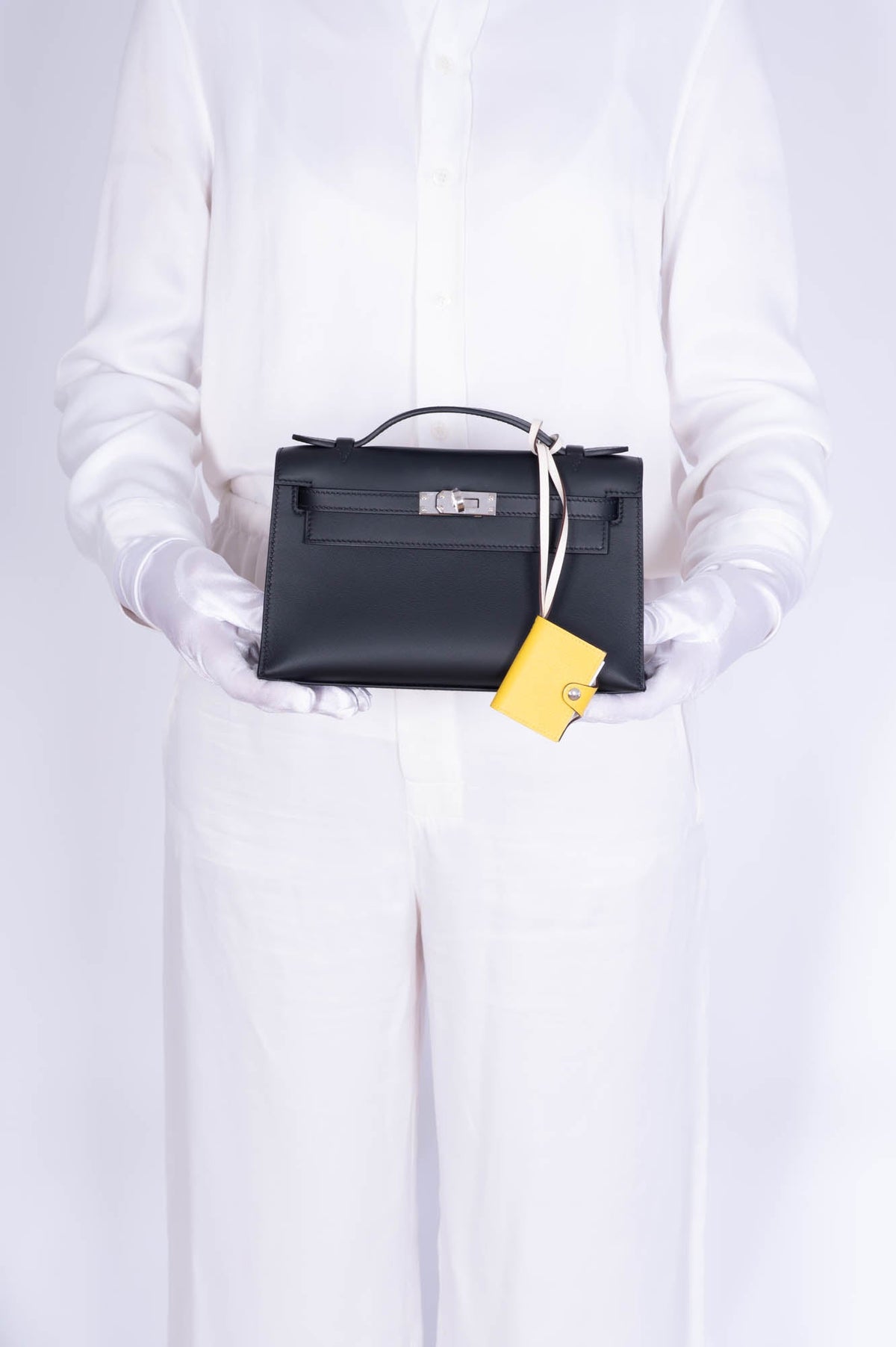 Hermès Kelly Pochette Noir Swift Palladium Hardware, held in both hands by a model in white gloves, styled with a charm, sold separately