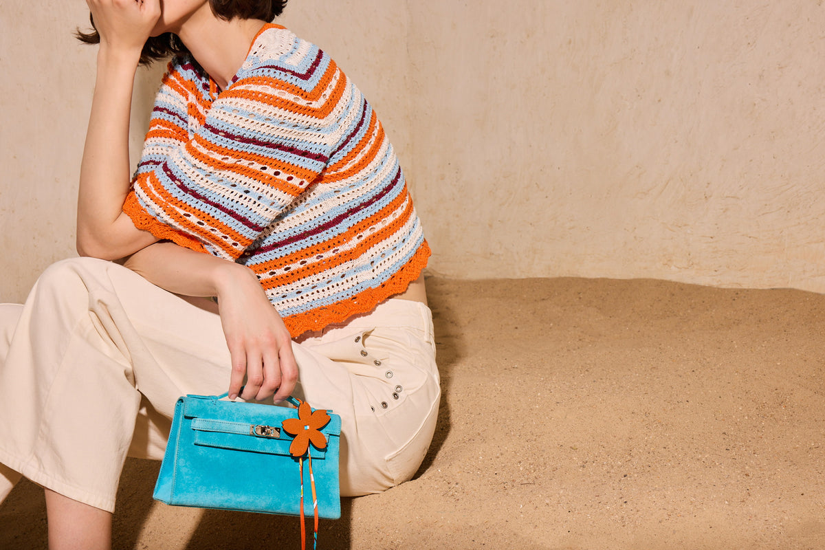 Hermès Kelly Pochette Turquoise Veau Doblis Suede Palladium Hardware, carried by a seated model, close view with a flower charm that is not included