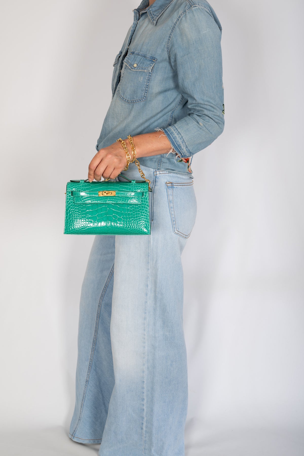 Hermès Kelly Pochette in Vert Jade shiny alligator with gold hardware, hand-carried by a model in denim.