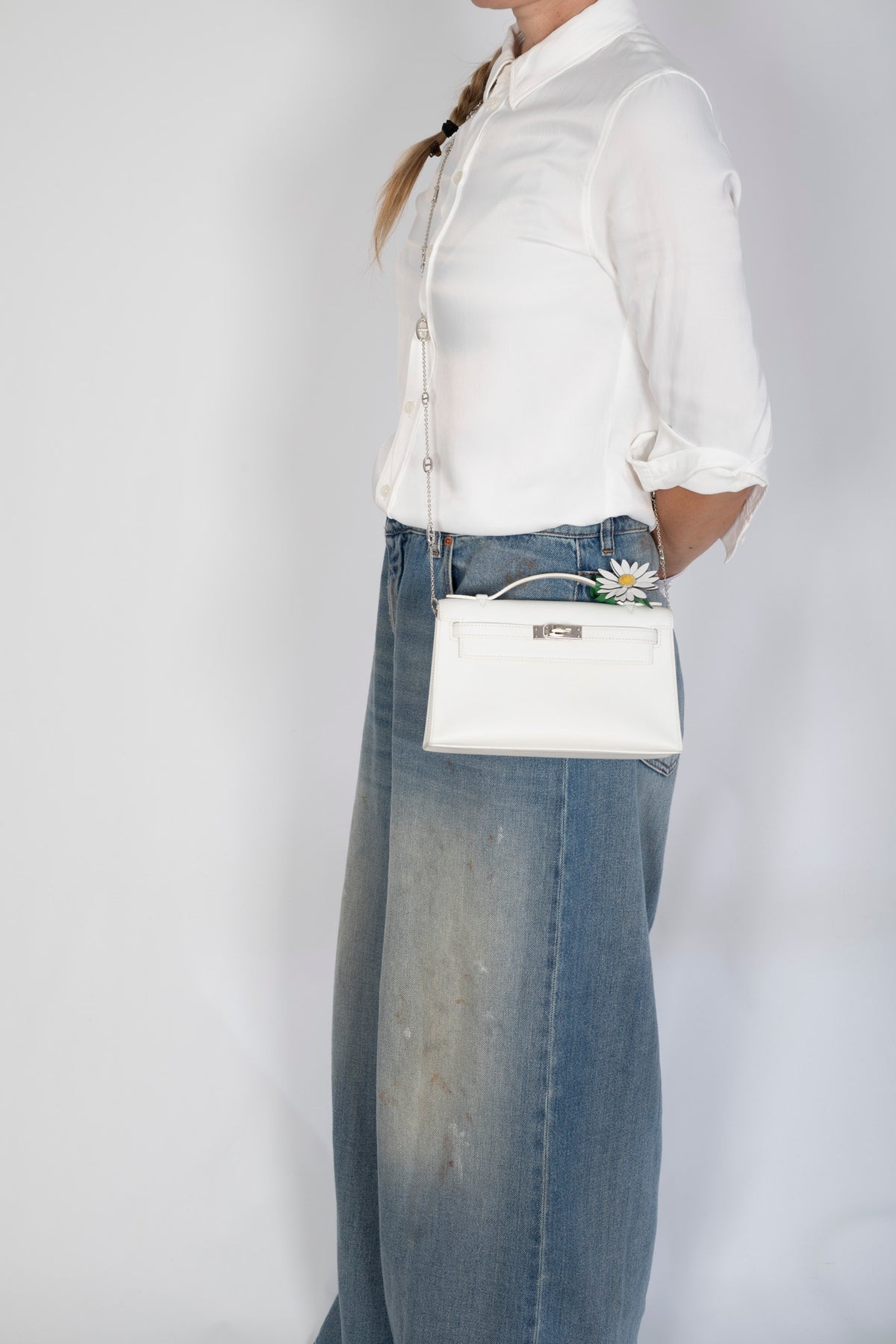 Hermès Kelly Pochette White Swift Palladium Hardware, held at the hip by a model, styled with a daisy charm, sold separately