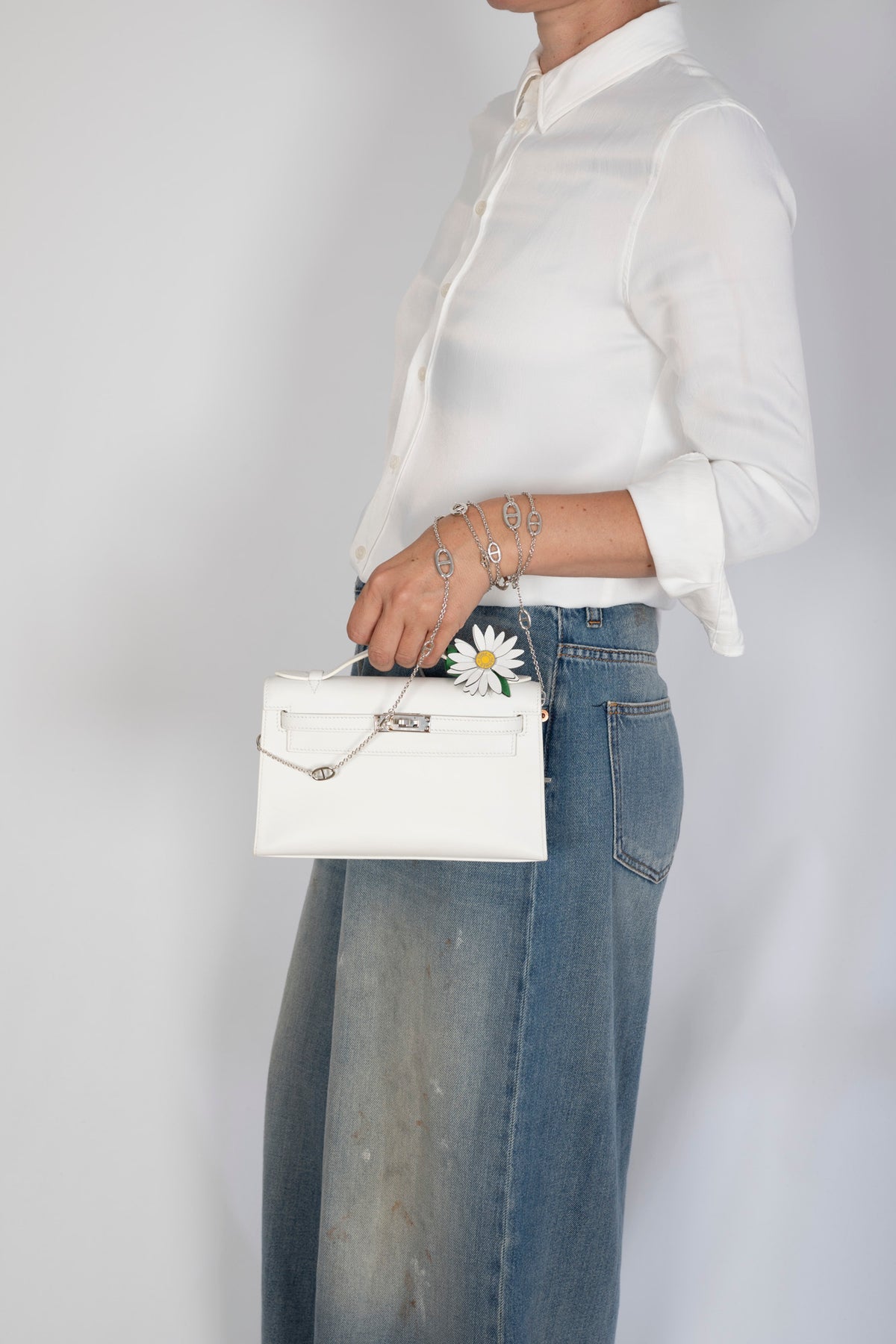 Hermès Kelly Pochette White Swift Palladium Hardware, held by its handle by a model, styled with a daisy charm, sold separately