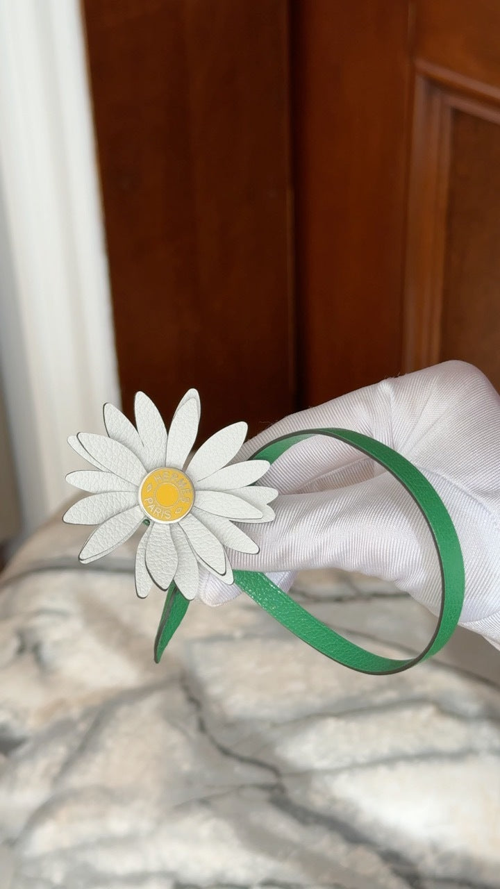 Hermès Marguerite Daisy Charm White Jaune de Naples Bambou, held in a gloved hand with the green strap curved