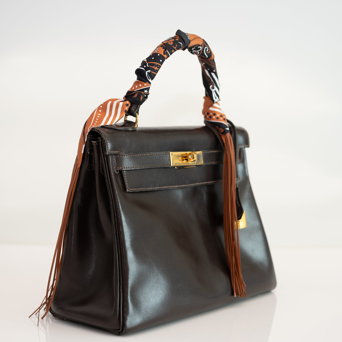 Hermès Vintage Kelly 28 Retourné Marron Foncé Box Calf Gold Hardware, three-quarter view, styled with a fringed twilly, sold separately