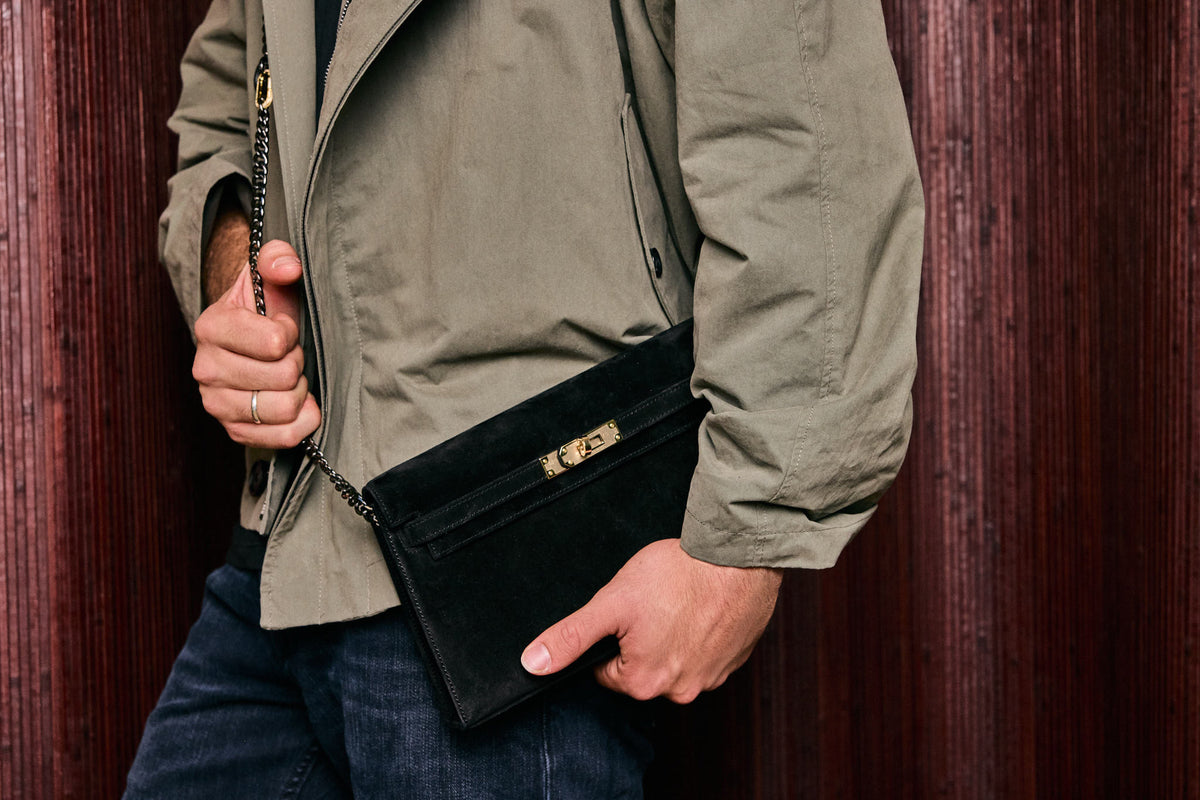Hermès Kelly Elan in Noir Veau Doblis suede with permabrass hardware, worn on a chain by a model in an olive jacket
