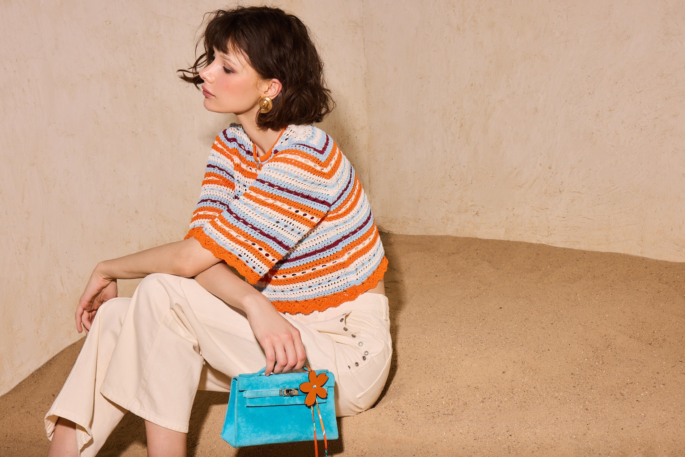 Model seated on sand holding a turquoise blue suede Mini Kelly with an orange flower charm — JaneFinds campaign